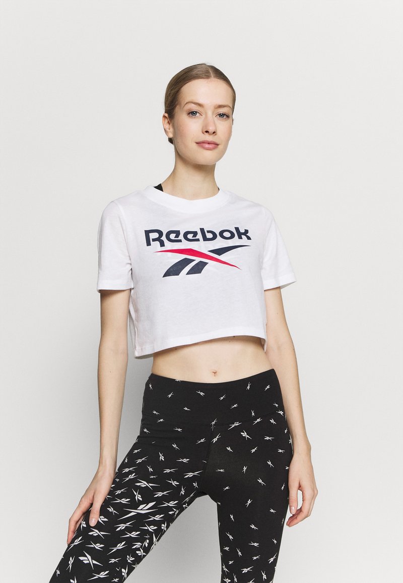 Weißes, verkürztes T-Shirt mit schwarzem und rotem Reebok-Logo, kombiniert mit schwarzen Leggings, die ein weißes abstraktes Muster und einen hohen Bund aufweisen.