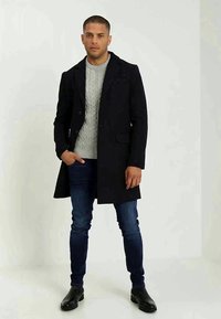 Abrigo de lana negro sobre un suéter de punto grueso gris claro, combinado con vaqueros azules y botas de cuero negras. De pie contra un fondo claro.
