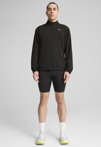 Jachetă ușoară de culoare neagră, cu guler înalt, fermoar frontal și manșete elastice, asociată cu pantaloni scurți negri mulați și adidași sport multicolori.