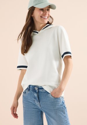 Femme souriante portant une casquette bleu clair, un haut blanc à manches courtes avec des rayures bleu marine sur les manches et le col, et un jean bleu clair, une main dans la poche.