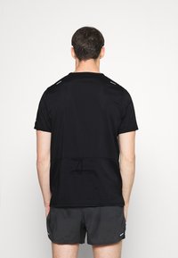 Černé sportovní tričko s krátkými rukávy, vybavené texturovanou tkaninou a reflexními detaily na ramenou, nošené s šedými šortkami.