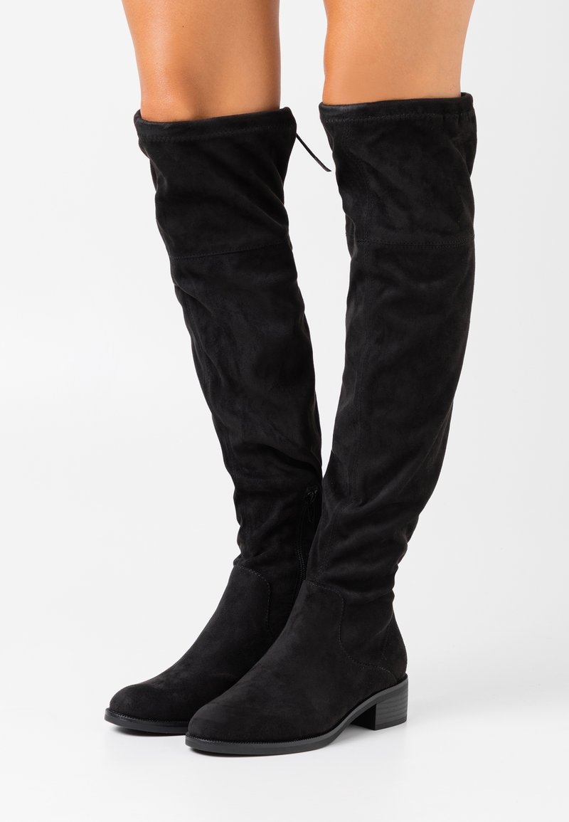 Bottes en daim noir montant au-dessus du genou avec un bout rond et un talon bloc. Les bottes présentent un design décontracté et une fermeture éclair latérale pour faciliter l'enfilage.