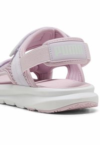 Gaiši rozā čības ar strukturētām auduma siksnām un mīkstu baltu zoli. Piekarē redzams izteikts Puma logo uz papēža siksnas.