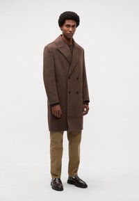 Cappotto di lana marrone doppiopetto con rever a scalpello, abbinato a pantaloni di corduroy color khaki e mocassini marroni scuri. Struttura liscia, vestibilità sartoriale.