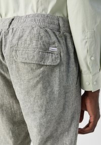 Gray linen pants with an elastic waistband, featuring a back pocket and a small brand label. Accompanied by a light green patterned shirt.