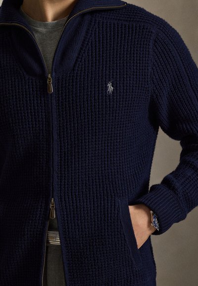 Navy knit zip-up cardigan with textured fabric, high collar, and subtle logo accent, featuring side pockets and ribbed cuffs.
