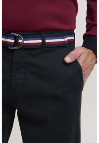 Black trousers featuring a multicolored woven belt with red, white, and blue stripes and a silver buckle, with a hand resting in the pocket.