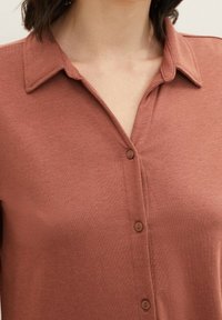 Nahaufnahme einer Person, die ein braunes Hemd mit Knöpfen und spitzem Kragen trägt, wobei der Halsausschnitt und der obere Brustbereich zu sehen sind.