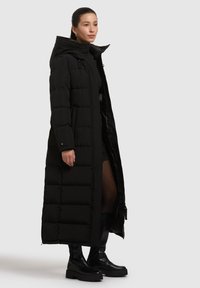 Schwarze Steppjacke mit langem Design, Reißverschluss und geräumiger Kapuze. Enthält ein gesteppte Muster und Seitentaschen. Getragen mit schwarzen kniehohen Stiefeln.
