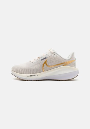 Buty do biegania Nike w jasnobeżowej tkaninie z teksturowaną cholewką, złotym logo Swoosh, białą środkową podeszwą oraz szarą gumową podeszwą z rowkami.