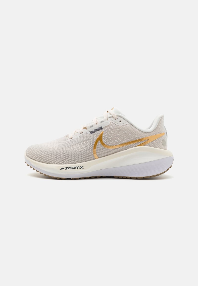 Nike hardloopschoen in lichtbeige stof met gestructureerd bovenwerk, gouden swoosh, witte tussenzool en een grijze rubberen buitenzool met groeven.