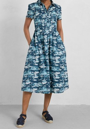 Femme portant une robe bleue à manches courtes avec col, imprimé nautique blanc, et des sandales fermées bleu marine avec des brides à la cheville.