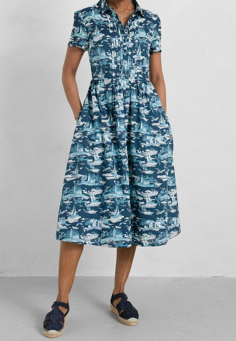 Femme portant une robe bleue à manches courtes avec col, imprimé nautique blanc, et des sandales fermées bleu marine avec des brides à la cheville.