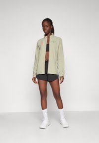 Light green, zippered athletic jacket with black logo, worn over a black crop top and paired with black shorts and white sneakers.