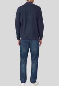 Marineblauwe gebreide trui met geribde kraag en achterkant, gecombineerd met een blauwe spijkerbroek. De outfit heeft een casual pasvorm en een gladde stoftextuur.