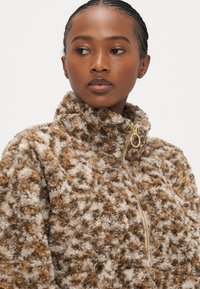 Bruine en crèmekleurige fleece trui met hoge kraag, halve rits en zachte, gestructureerde oppervlakte. Heeft een gouden ritssluiting detail.