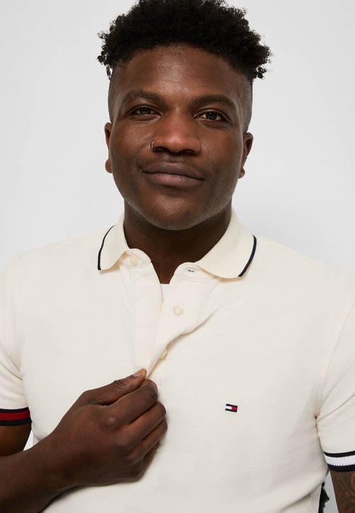 Krémová polo košile s texturovanou tkaninou, se zapínáním na tři knoflíky a malým vyšitým logem. Podtržená námořnickými a červenými pruhem na rukávech.