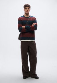 Πολύχρωμο ριγέ πουκάμισο με μακριά μανίκια σε βυσσινί και ναυτικό μπλε, συνδυασμένο με καφέ παντελόνια με ευρύ πόδι και μαύρα παπούτσια. Φαίνεται μια τσάντα ώμου.