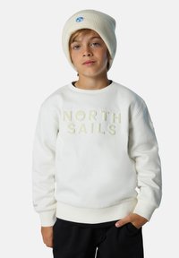 Sudadera blanca con "NORTH SAILS" en letras en relieve, combinada con un gorro de canalé crema. Pantalones negros lisos completan el conjunto.