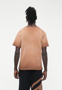 Vyras su juodomis dreadlock plaukais, dėvintis rudą trumpomis rankovėmis marškinį ir juodas kelnes su rudu abstrakčiu raštu, stovintis nugara.