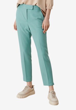 Pantalons tailleur bleu-vert clair avec une texture lisse, présentant des plis avant et des poches latérales, associés à des baskets épaisses beiges.