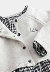 Jasnoszary, dziany kardigan z ciemnymi guzikami i haftem "hello"; posiada granatowo-białą kratkę w tle, miękka struktura.
