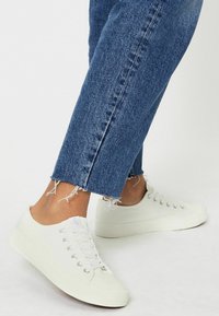 Baskets en toile blanches avec bout rond, fermeture à lacets, œillets en métal et semelle en caoutchouc texturée, associées à un jean bleu avec ourlet effiloché.
