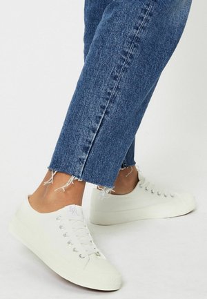 Baskets en toile blanches avec bout rond, fermeture à lacets, œillets en métal et semelle en caoutchouc texturée, associées à un jean bleu avec ourlet effiloché.