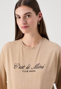 Bežs liela izmēra T-krekls ar apaļu kaklu un īsām piedurknēm, uz kura ir melna izšūta teksts "C'est le Mini CLUB MAMA." Gluda tekstūra.