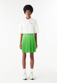 Camisa polo blanca de manga corta, falda plisada verde, con un logo de Lacoste en la cinturilla, combinada con calcetines blancos y zapatillas.