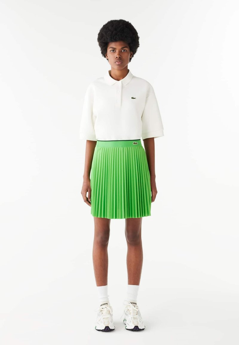Camisa polo blanca de manga corta, falda plisada verde, con un logo de Lacoste en la cinturilla, combinada con calcetines blancos y zapatillas.