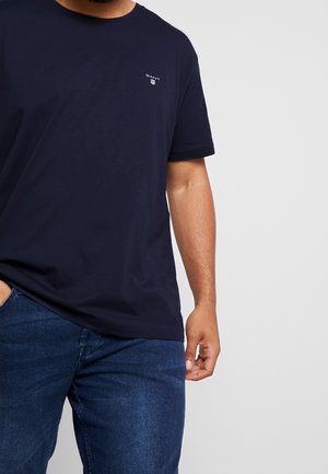 Homme portant un t-shirt à manches courtes GANT bleu marine et un jean en denim bleu, une main dans la poche et l'autre détendue le long du corps.