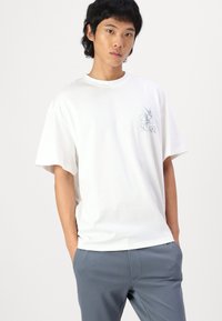 Camiseta de algodón blanca de manga corta, con un sutil estampado gráfico en azul claro en el lado izquierdo del pecho. Corte relajado con cuello redondo.