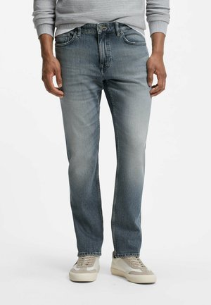 Uomo che indossa jeans blu scoloriti, maglione grigio e sneakers bianche, in piedi con le mani rilassate lungo i fianchi.
