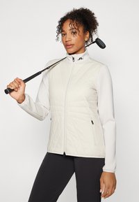 Lätt, krämfärgad quiltad väst med hög krage, dragkedjestängning och sidofickor, som bärs över en vit långärmad topp, hålls med en golfklubba.