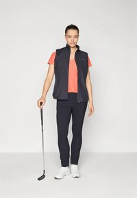 Veste sans manches bleu marine portée sur une chemise orange, assortie à un pantalon bleu marine et des baskets blanches. Un club de golf est tenu dans la main gauche.