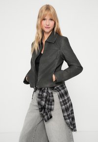Veste en faux cuir gris foncé avec col, poches avant et fermeture éclair ; associée à un pantalon large gris clair et une chemise à carreaux noir et blanc nouée à la taille.