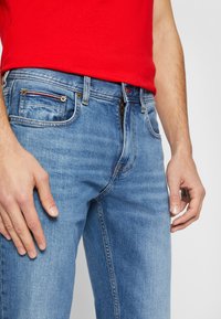 Jeans en denim bleu clair avec une fermeture éclair, design à cinq poches et détails de coutures rouges contrastants. Porté avec une chemise rouge.
