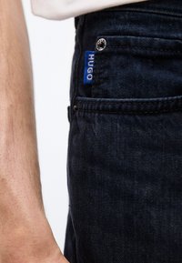 Κοντινό πλάνο τσέπης σκούρου τζιν με μπλε ετικέτα "HUGO" και νίκελ, δίπλα στο χέρι ενός ατόμου που φοράει ανοιχτόχρωμο πουκάμισο.