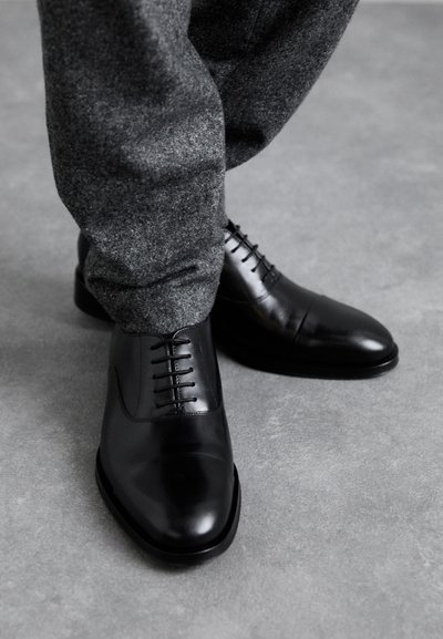 Scarpe eleganti in pelle nera con finitura lucida, dotate di punta arrotondata e lacci, abbinate a pantaloni grigi con texture.