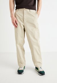 Beige cotton pants with a straight fit and button closure. Paired with green and yellow sneakers featuring a textured sole.
