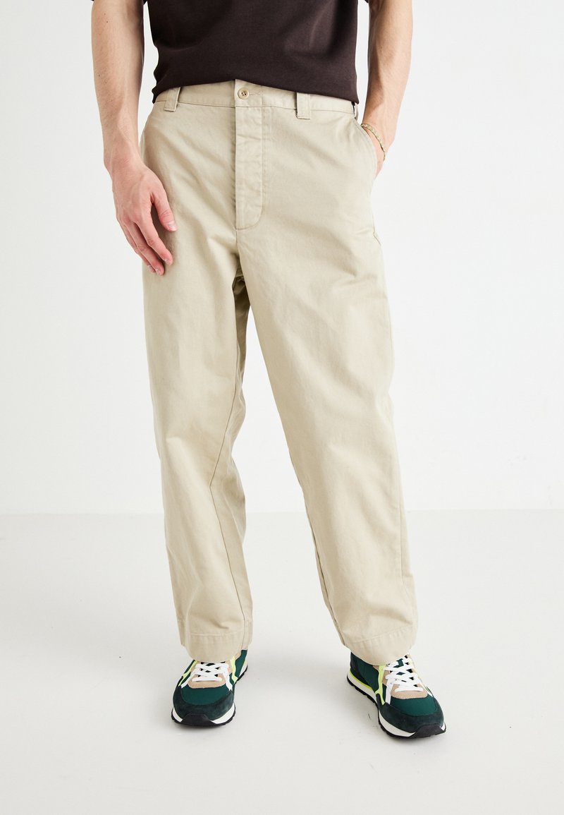Beige cotton pants with a straight fit and button closure. Paired with green and yellow sneakers featuring a textured sole.