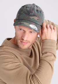 Căciulă de camuflaj cu visieră curbă și curea ajustabilă, confecționată dintr-un material cu un model texturat, combinând nuanțe de verde și maro.