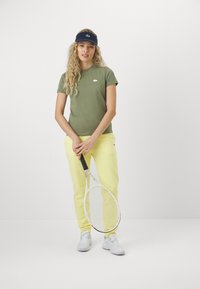 Oliven grønn tettsittende t-skjorte, gule sweatpants og marineblå caps; holder en tenniskølle. Enkelt design, minimal merkevare på klærne.