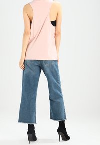 Kvinna i ärmlös rosa topp, cropped blå jeans med fransiga kanter och svarta högklackade stövlar, står vänd bort från kameran.