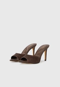 Une paire de mules ouvertes à bout en cuir suédé marron foncé avec des talons aiguilles fins sur un fond clair.