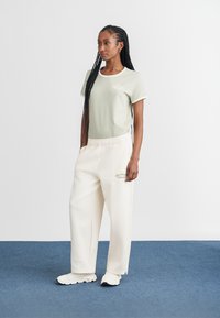 Light green t-shirt with white trim and logo; paired with loose white pants featuring green embroidery; worn with white sneakers.