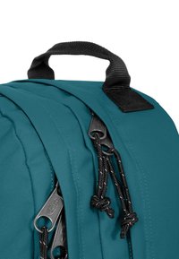 Tyrkysový batoh s texturovanou látkou, horní rukojetí a kontrastními černými zipy a šňůrkami pro přístup k oddělením.