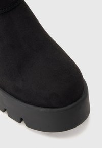 Gros plan sur l'embout d'une botte en daim noir avec une semelle en caoutchouc épaisse et texturée sur un fond blanc uni.