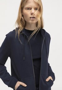 Marinblå zip-up hoodie med en mjuk textur, som har en frontficka, snoddar och ribbade ärmslut, bärs över en matchande topp.